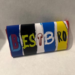 Puede incluir: Una envoltura rectangular de barra de chocolate con un diseño de papel rasgado con rayas verticales en azul, negro, blanco, azul y amarillo. El texto BEST BRO está impreso en el frente con letras estilizadas, audaces y multicolores.