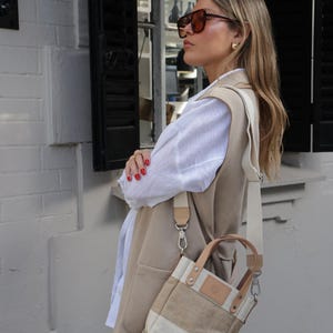 May include: A woman wearing a beige vest over a white shirt and blue jeans. She is carrying a beige and white canvas tote bag with a brown leather strap.