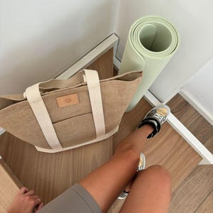 Op de afbeelding: Een bruine tote bag met cr&egrave;mekleurige bandjes en een klein leren label, een opgerolde lichtgroene yogamat en grijze shorts. De tas staat op een houten oppervlak naast een witte muur.