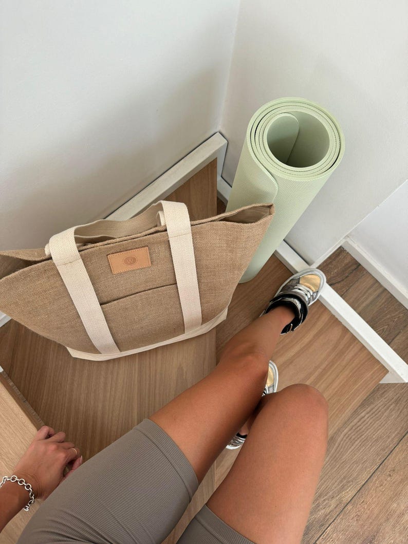 Op de afbeelding: Een bruine tote bag met cr&egrave;mekleurige bandjes en een klein leren label, een opgerolde lichtgroene yogamat en grijze shorts. De tas staat op een houten oppervlak naast een witte muur.
