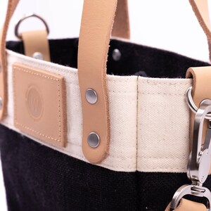 May include: Close-up of a black and white canvas tote bag with brown leather handles and a metal clasp. The bag has a small brown leather patch with a logo on the front.