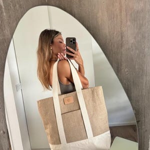 Op de afbeelding: Een grote draagtas in natuurlijk beige met een witte basis. Het heeft witte banden en een kleine, rechthoekige leren patch. De tas is gemaakt van geweven materiaal en heeft een voorvak. Ideaal om te winkelen of naar het strand te gaan.