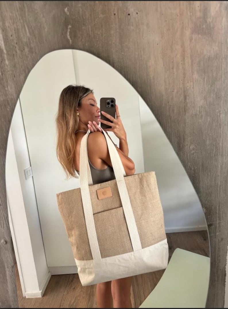 Op de afbeelding: Een grote draagtas in natuurlijk beige met een witte basis. Het heeft witte banden en een kleine, rechthoekige leren patch. De tas is gemaakt van geweven materiaal en heeft een voorvak. Ideaal om te winkelen of naar het strand te gaan.
