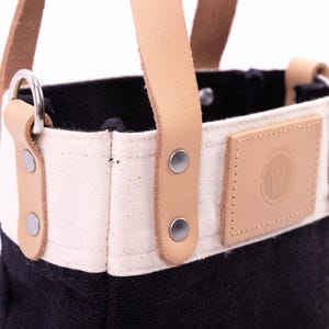 May include: A small black and white canvas tote bag with leather handles and a leather patch with the letter "W" in a circle. The bag has metal rivets on the handles.