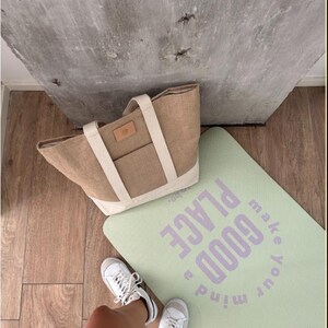 Op de afbeelding: Een lichtbruine tote bag met cr&egrave;mekleurige bandjes en een klein voorvak ligt naast een lichtgroene yogamat. Op de yogamat staat in paarse letters "make your mind a GOOD PLACE". Witte sneakers zijn zichtbaar.