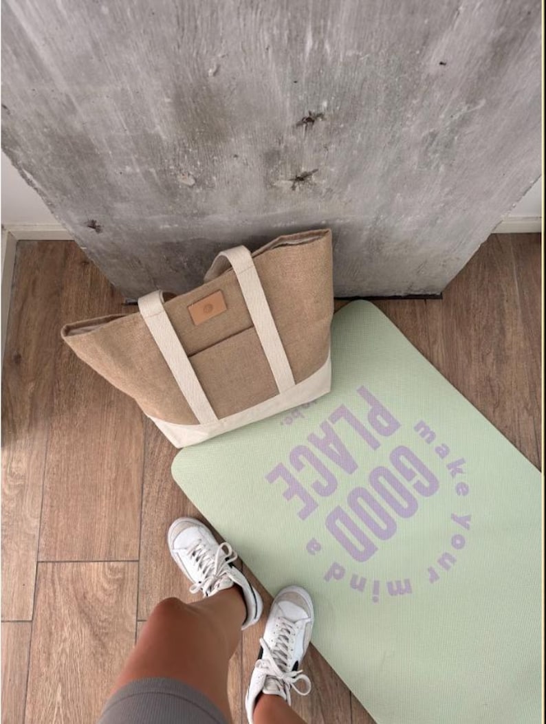 Op de afbeelding: Een lichtbruine tote bag met cr&egrave;mekleurige bandjes en een klein voorvak ligt naast een lichtgroene yogamat. Op de yogamat staat in paarse letters "make your mind a GOOD PLACE". Witte sneakers zijn zichtbaar.