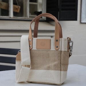 May include: A small, beige and white canvas tote bag with brown leather handles and a shoulder strap. The bag has a small, round leather patch with a logo on the front.