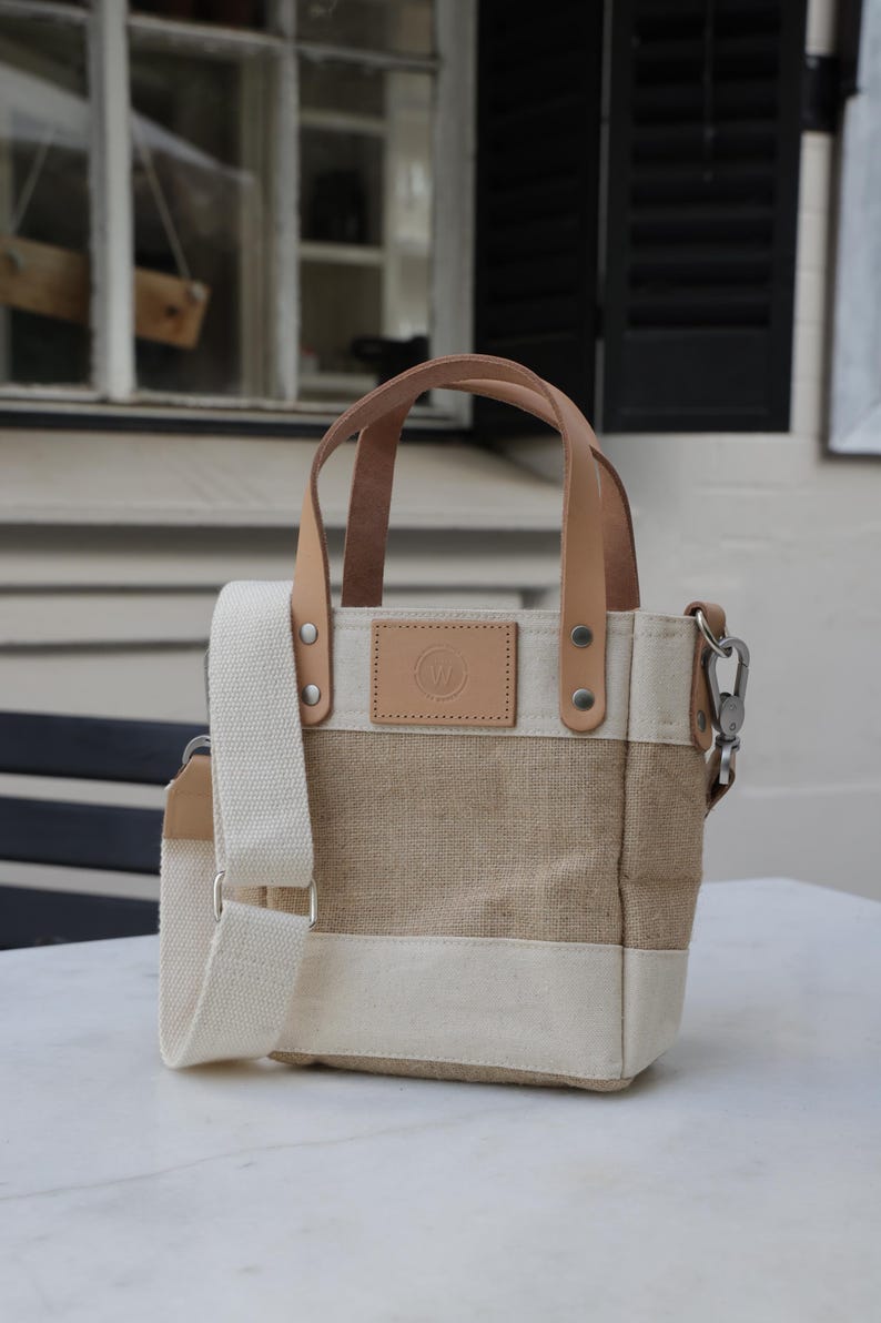 May include: A small, beige and white canvas tote bag with brown leather handles and a shoulder strap. The bag has a small, round leather patch with a logo on the front.