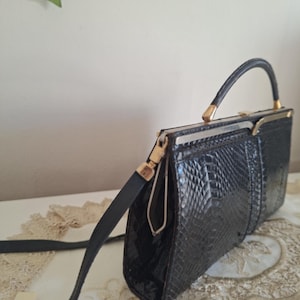 Puede incluir: Bolso de mano vintage negro con un patrón texturizado similar al de un reptil. El bolso tiene un asa superior y una correa larga y desmontable para el hombro. Se ven detalles metálicos dorados en el marco y el cierre. El bolso está sobre una superficie blanca.