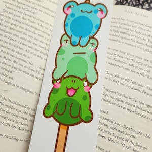 May include: A whimsical bookmark featuring a stack of three cartoon frogs on a popsicle stick. The frogs are in shades of blue and green with pink accents. The bookmark is rectangular and printed on white cardstock.
