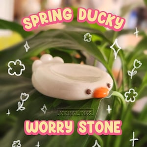 Puede incluir: Una piedra de preocupación de cerámica blanca con forma de pato, con un cuerpo liso y redondeado, dos pequeños ojos marrones y un pico naranja brillante. El texto SPRING DUCKY WORRY STONE aparece en rosa y amarillo, con flores dibujadas.