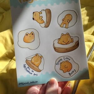 Könnte beinhalten: Eine Bogen mit Aufklebern, die Comic-Frösche zeigen, die als Eggs Benedict verkleidet sind. Die Aufkleber haben verschiedene Sätze, darunter "It's an Eggcellent Day" und "Sleepy Little Baby".