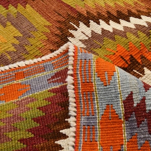 May include: Close-up of a colourful woven rug with a geometric pattern. The rug features a mix of orange, brown, green, and purple hues. The edges are finished with a white fringe.