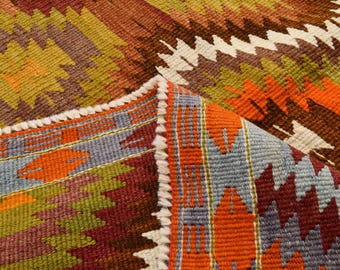 Può includere: Primo piano di un tappeto tessuto colorato con un motivo geometrico. Il tappeto presenta un mix di tonalità arancioni, marroni, verdi e viola. I bordi sono rifiniti con una frangia bianca.
