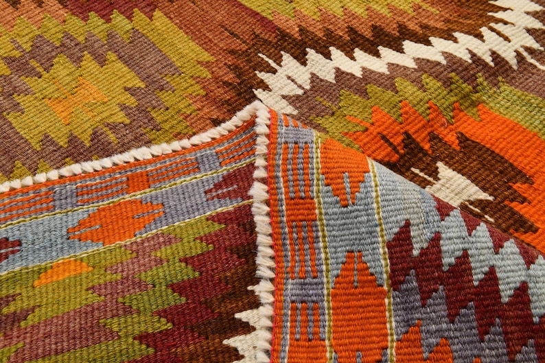 May include: Close-up of a colourful woven rug with a geometric pattern. The rug features a mix of orange, brown, green, and purple hues. The edges are finished with a white fringe.