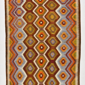 May include: A rectangular rug with a geometric diamond pattern in shades of brown, orange, green, grey, and white. The rug measures 290 x 177 cm. The edges are bordered with a dark brown trim. The rug has a woven texture.
