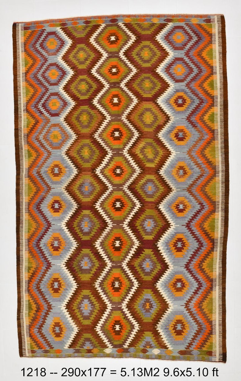 May include: A rectangular rug with a geometric diamond pattern in shades of brown, orange, green, grey, and white. The rug measures 290 x 177 cm. The edges are bordered with a dark brown trim. The rug has a woven texture.