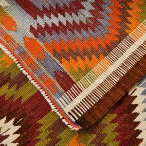 May include: Close-up of a colourful, patterned rug with a geometric design. The rug features a combination of colours including red, orange, green, and blue. The edges are finished with a white and brown border.