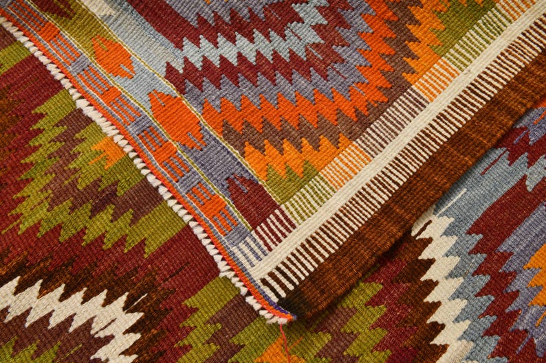May include: Close-up of a colourful, patterned rug with a geometric design. The rug features a combination of colours including red, orange, green, and blue. The edges are finished with a white and brown border.