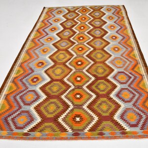 May include: Rectangular rug featuring a geometric diamond pattern in shades of brown, orange, yellow, and grey. The rug has a brown border and a repeating diamond design. The rug is placed on a white surface.