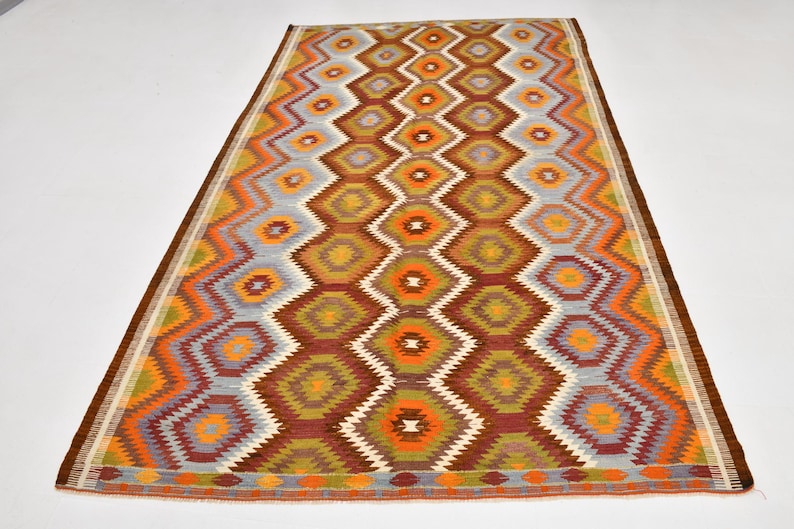 May include: Rectangular rug featuring a geometric diamond pattern in shades of brown, orange, yellow, and grey. The rug has a brown border and a repeating diamond design. The rug is placed on a white surface.
