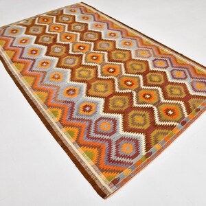 May include: Rectangular rug with a geometric pattern in shades of brown, orange, yellow, and blue. The rug features a repeating diamond and zigzag design, with a brown border. The rug is likely made of woven material.