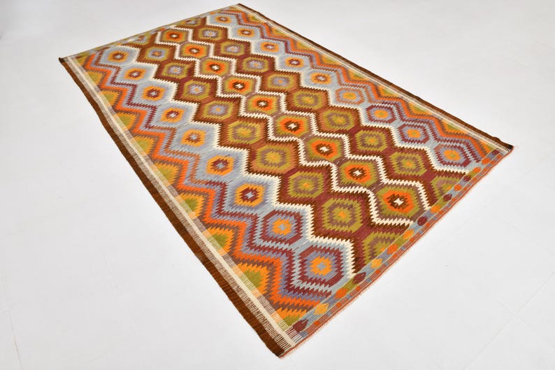 May include: Rectangular rug with a geometric pattern in shades of brown, orange, yellow, and blue. The rug features a repeating diamond and zigzag design, with a brown border. The rug is likely made of woven material.