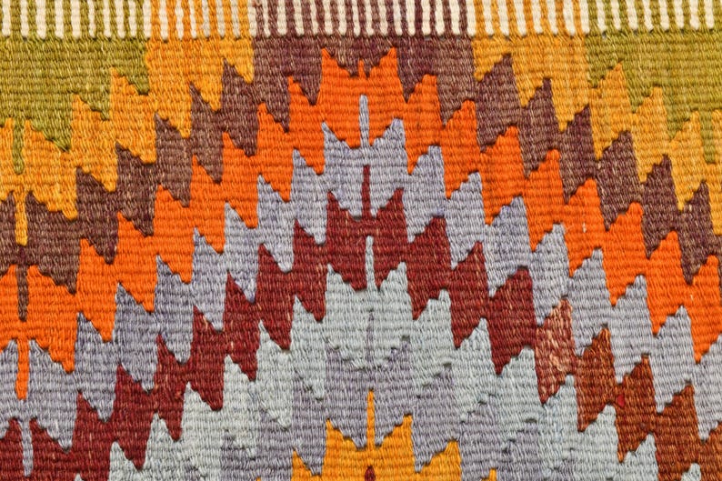 May include: Close-up of a woven textile with a repeating geometric pattern. The design features zig-zag shapes in shades of orange, red, grey, and brown. The top edge has a striped border with green, yellow, and white. This textile could be a rug or wall hanging.