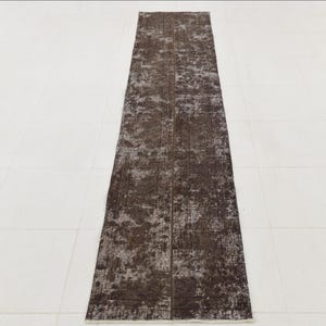 May include: A long, rectangular rug with a distressed, vintage appearance. The rug features a dark brown colour with lighter, faded areas, creating a mottled effect. The rug is placed on a white tiled floor.