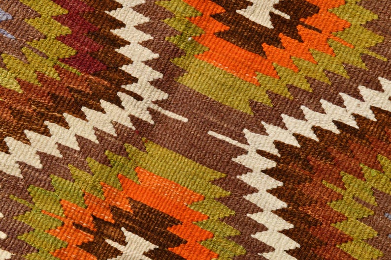May include: Close-up of a woven textile with a repeating geometric pattern. The design features zig-zag shapes in shades of brown, orange, green, and white. The textile has a textured appearance, suggesting a rug or fabric.