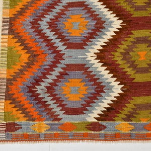 May include: Hand-woven rug with a geometric pattern in shades of orange, red, green, blue, and white. The rug features a repeating diamond design with a brown border. A colourful and textured floor covering.