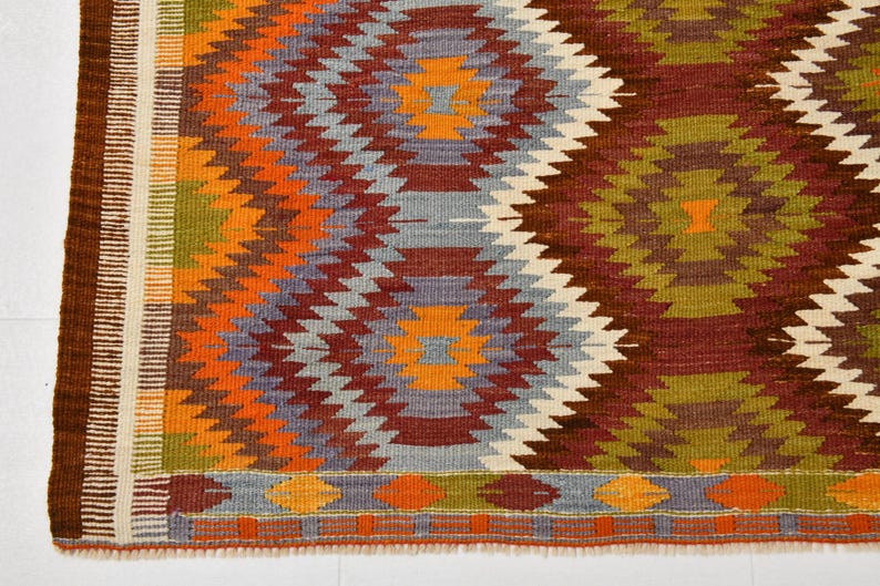 May include: Hand-woven rug with a geometric pattern in shades of orange, red, green, blue, and white. The rug features a repeating diamond design with a brown border. A colourful and textured floor covering.