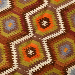 May include: Close-up of a woven rug with a repeating geometric pattern. The design features diamond shapes in shades of brown, orange, green, and blue, outlined with white zigzags. The rug has a textured appearance.