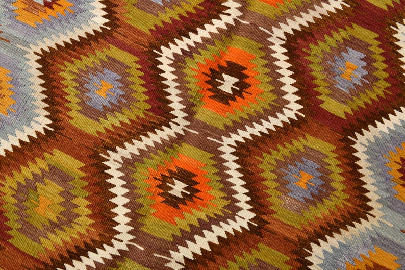 May include: Close-up of a woven rug with a repeating geometric pattern. The design features diamond shapes in shades of brown, orange, green, and blue, outlined with white zigzags. The rug has a textured appearance.