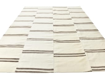 Op de afbeelding: Een rechthoekig vloerkleed met een crèmekleurige basis en herhalende horizontale donkerbruine strepen. Het kleed is samengesteld uit meerdere verticaal aan elkaar genaaide panelen, wat een consistent patroon van dunne, donkerbruine lijnen vormt.