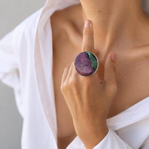 Puede incluir: Un anillo grande y redondo con una banda plateada. El anillo presenta una piedra pulida con una mezcla de colores rojo oscuro y verde. El anillo se lleva en un dedo, sobre una camisa blanca.