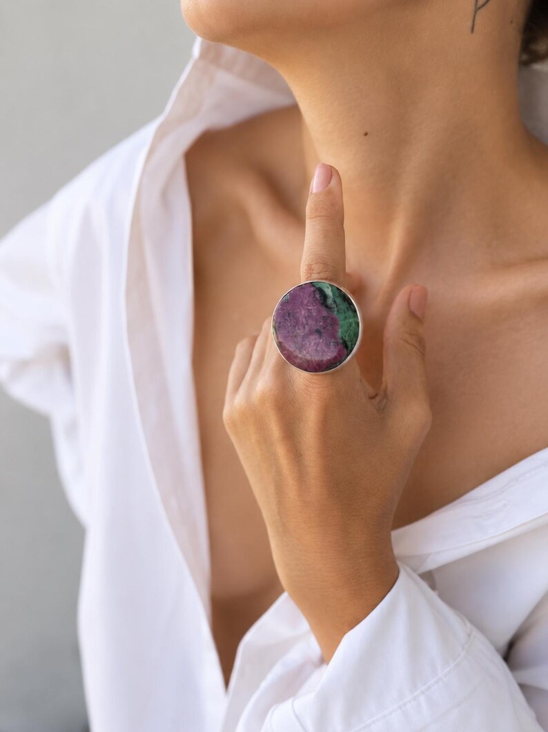 Puede incluir: Un anillo grande y redondo con una banda plateada. El anillo presenta una piedra pulida con una mezcla de colores rojo oscuro y verde. El anillo se lleva en un dedo, sobre una camisa blanca.