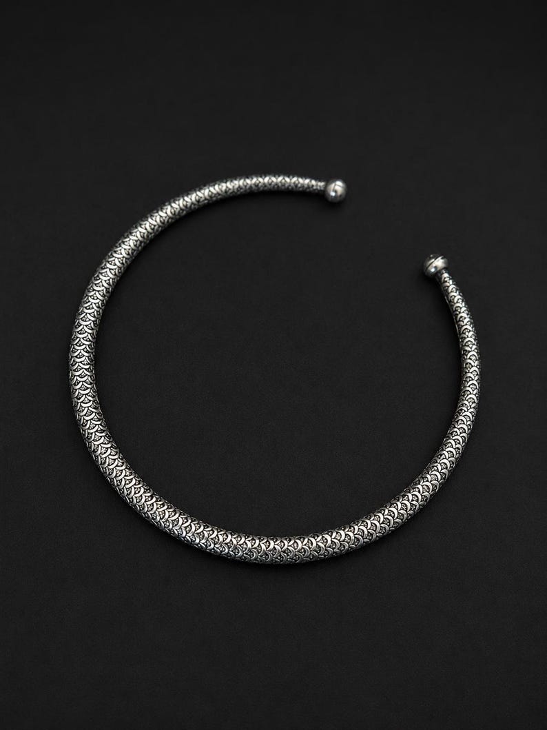 Peut inclure: Un collier ras du cou argent&eacute; avec un motif textur&eacute; en forme d'&eacute;cailles. Le design ouvert pr&eacute;sente une petite boule &agrave; chaque extr&eacute;mit&eacute;. Le collier est pos&eacute; sur un fond sombre, soulignant ses d&eacute;tails complexes et son &eacute;clat m&eacute;tallique.