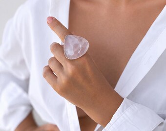 Può includere: Un grande anello in quarzo rosa chiaro con montatura argentata. La gemma triangolare è lucidata e riflette la luce. L'anello è indossato su un dito, su una camicia bianca.