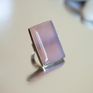Pu&ograve; includere: Un anello con una pietra rettangolare liscia di colore rosa pallido incastonata in una montatura in metallo color argento lucido. La pietra ha un aspetto morbido e traslucido con una lucentezza sottile, e la fascia in metallo &egrave; visibile.