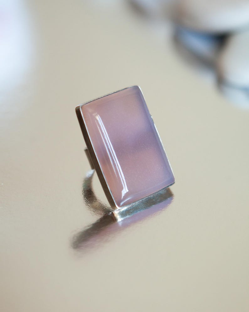 Pu&ograve; includere: Un anello con una pietra rettangolare liscia di colore rosa pallido incastonata in una montatura in metallo color argento lucido. La pietra ha un aspetto morbido e traslucido con una lucentezza sottile, e la fascia in metallo &egrave; visibile.