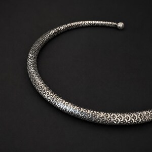 Peut inclure: Un collier ras du cou argent&eacute; et textur&eacute; avec un motif r&eacute;p&eacute;titif. Le collier forme un demi-cercle, avec une petite boule &agrave; l'extr&eacute;mit&eacute;. Le fond est noir uni.