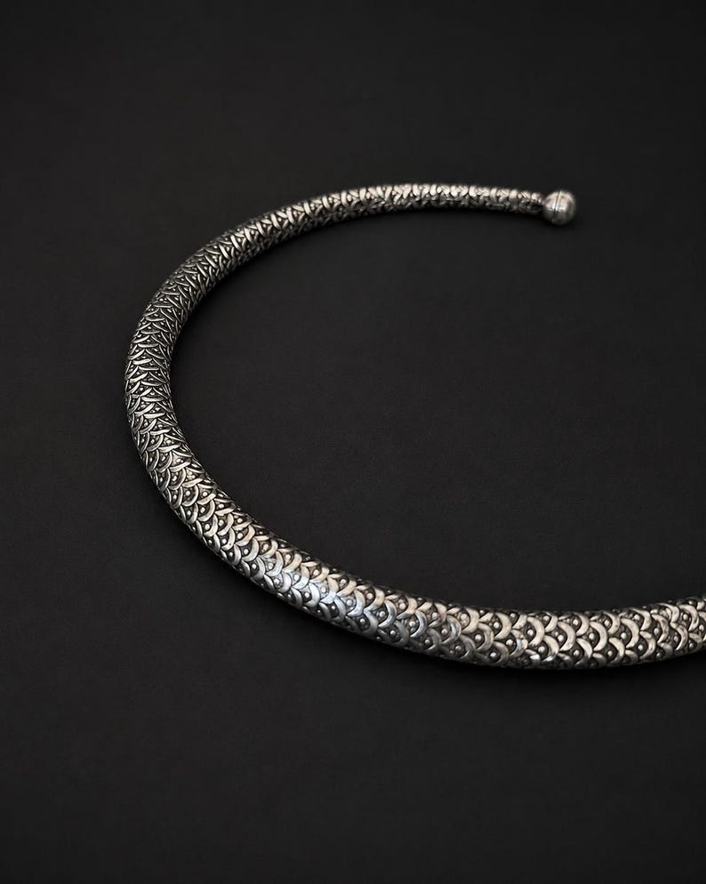 Peut inclure: Un collier ras du cou argent&eacute; et textur&eacute; avec un motif r&eacute;p&eacute;titif. Le collier forme un demi-cercle, avec une petite boule &agrave; l'extr&eacute;mit&eacute;. Le fond est noir uni.