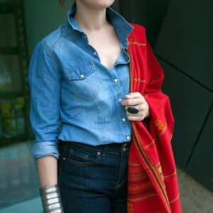 May include: A woman wearing a light blue denim shirt, dark blue jeans, and a red shawl with an intricate pattern. She is also wearing a silver cuff bracelet and a large black ring. The outfit is casual and stylish.