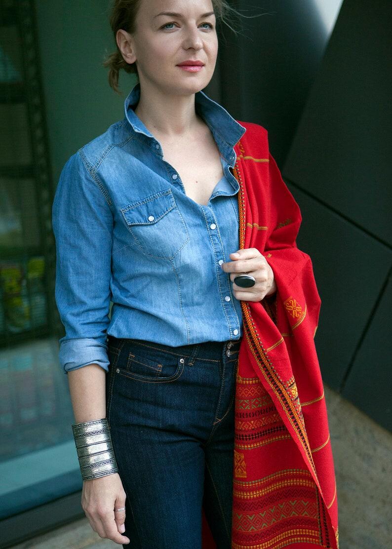 May include: A woman wearing a light blue denim shirt, dark blue jeans, and a red shawl with an intricate pattern. She is also wearing a silver cuff bracelet and a large black ring. The outfit is casual and stylish.