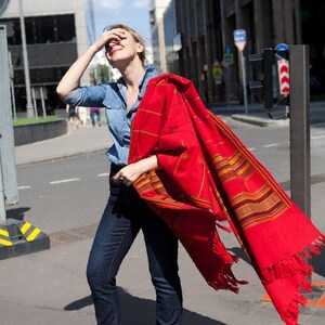 May include: A vibrant red shawl with a geometric pattern and fringe detail. The shawl is held by a person wearing a denim shirt and dark blue jeans. The shawl's design includes gold and brown stripes.