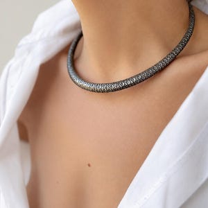 Peut inclure: Un collier argent&eacute; avec un motif r&eacute;p&eacute;titif de formes incurv&eacute;es. Le collier est port&eacute; sur une chemise blanche. Le design est de style ras du cou, pr&egrave;s du cou, et semble &ecirc;tre en m&eacute;tal.