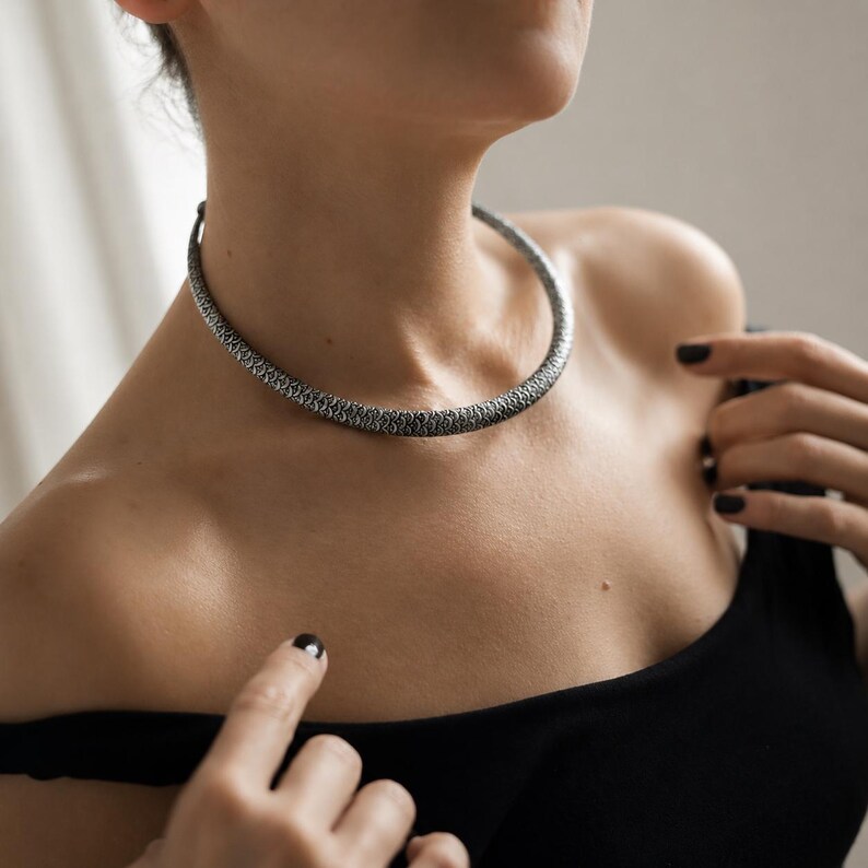 Peut inclure: Un collier ras du cou argent&eacute; avec un motif textur&eacute; et tiss&eacute;. Le collier est port&eacute; autour du cou. Le mod&egrave;le porte un haut noir. Le collier mesure environ 40 cm de long.
