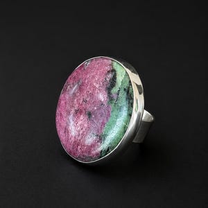 Puede incluir: Anillo llamativo con una piedra preciosa ovalada pulida en tonos rosa, verde y negro, engastada en una ancha banda de plata pulida. El anillo se muestra sobre un fondo negro. Una joya audaz.