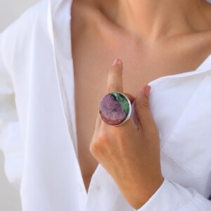 Puede incluir: Un anillo grande y redondo con una banda plateada. El anillo presenta una piedra natural con una mezcla de colores rosa, verde y blanco. El anillo se lleva en un dedo, y la persona lleva una camisa blanca abotonada.